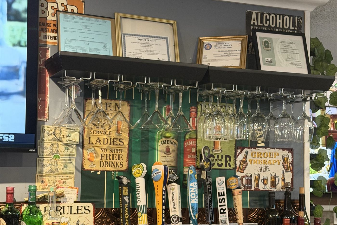 Vintage bar signs and framed licences above a rack of hanging glassware.